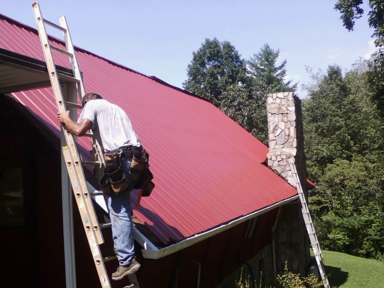 Roofer on a ladder beside a red metal roof and stone chimney — Soffit & Fascia Repair by Roofing Brothers in Pomona Park